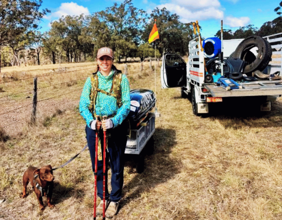 Woman in teal jacket with trekking poles beside a small brown dog in a dry field; gear trailer and truck in background.