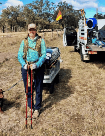 Woman in teal jacket with trekking poles beside a small brown dog in a dry field; gear trailer and truck in background.