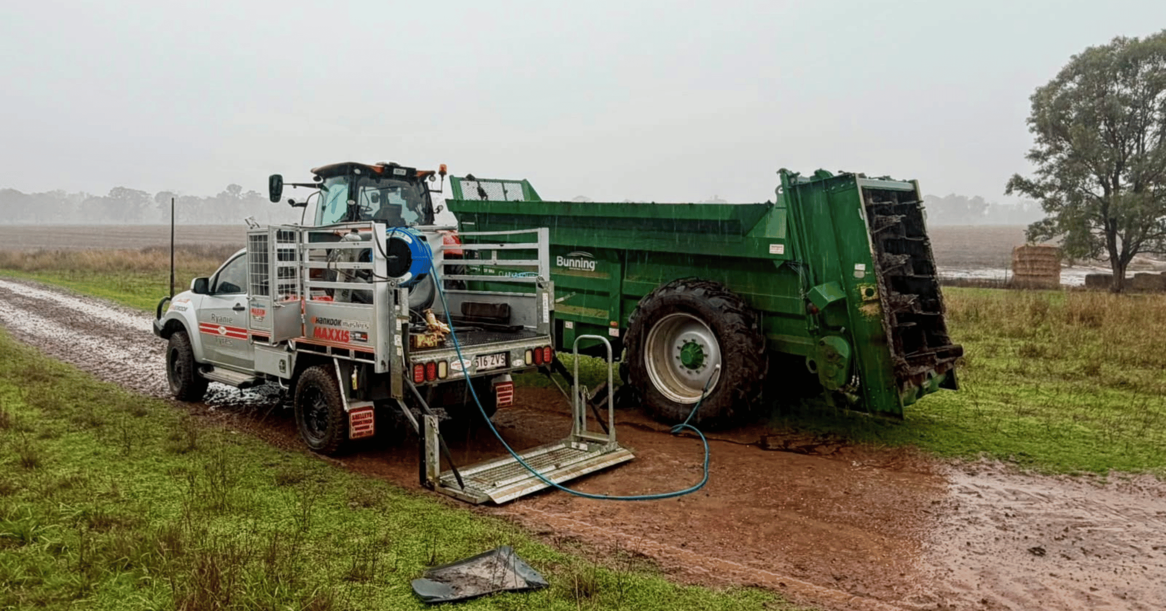 White recovery truck attached to a large green dump trailer stuck in a muddy rural field.