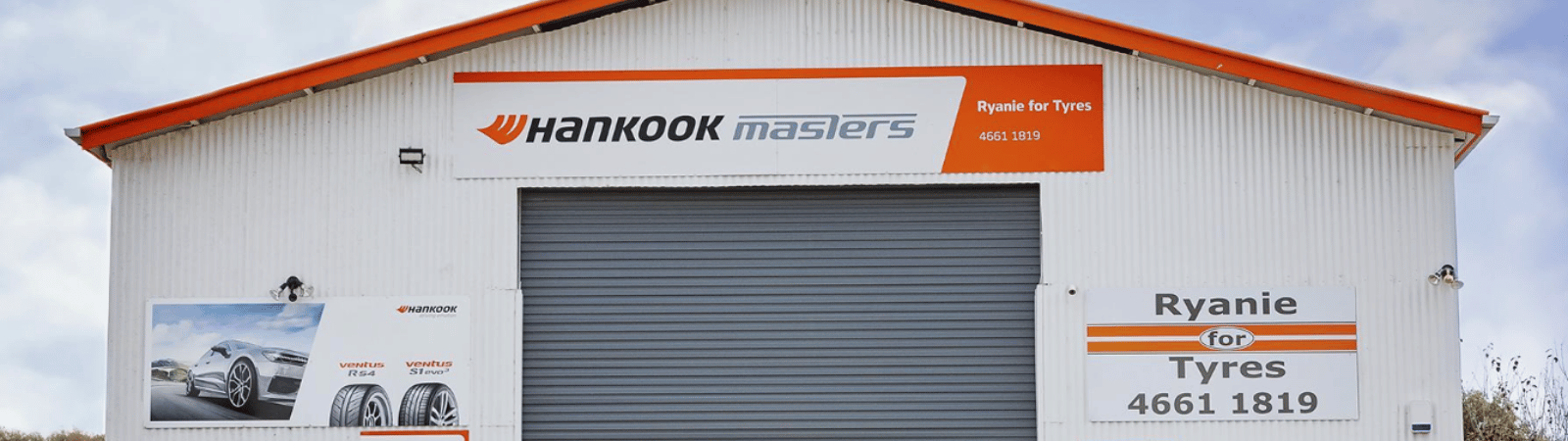 Tire shop front with a large gray roll-door, white corrugated walls, orange roof trim, and signs reading Hankook Masters and Ryanie for Tyres, 4661 1819.