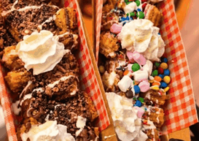 Two dessert trays in red-checkered boxes held by hands, piled with cake pieces, whipped cream, and colorful candies and sprinkles in a festive presentation.