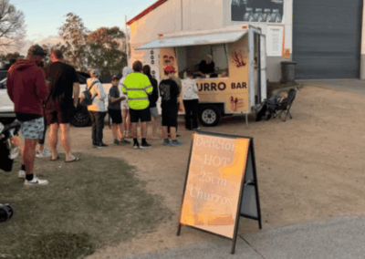 People line up at a small food truck labeled 'BURRO BAR' outside a warehouse, taking orders from a server. A bright sign advertises hot chimis/churros nearby.